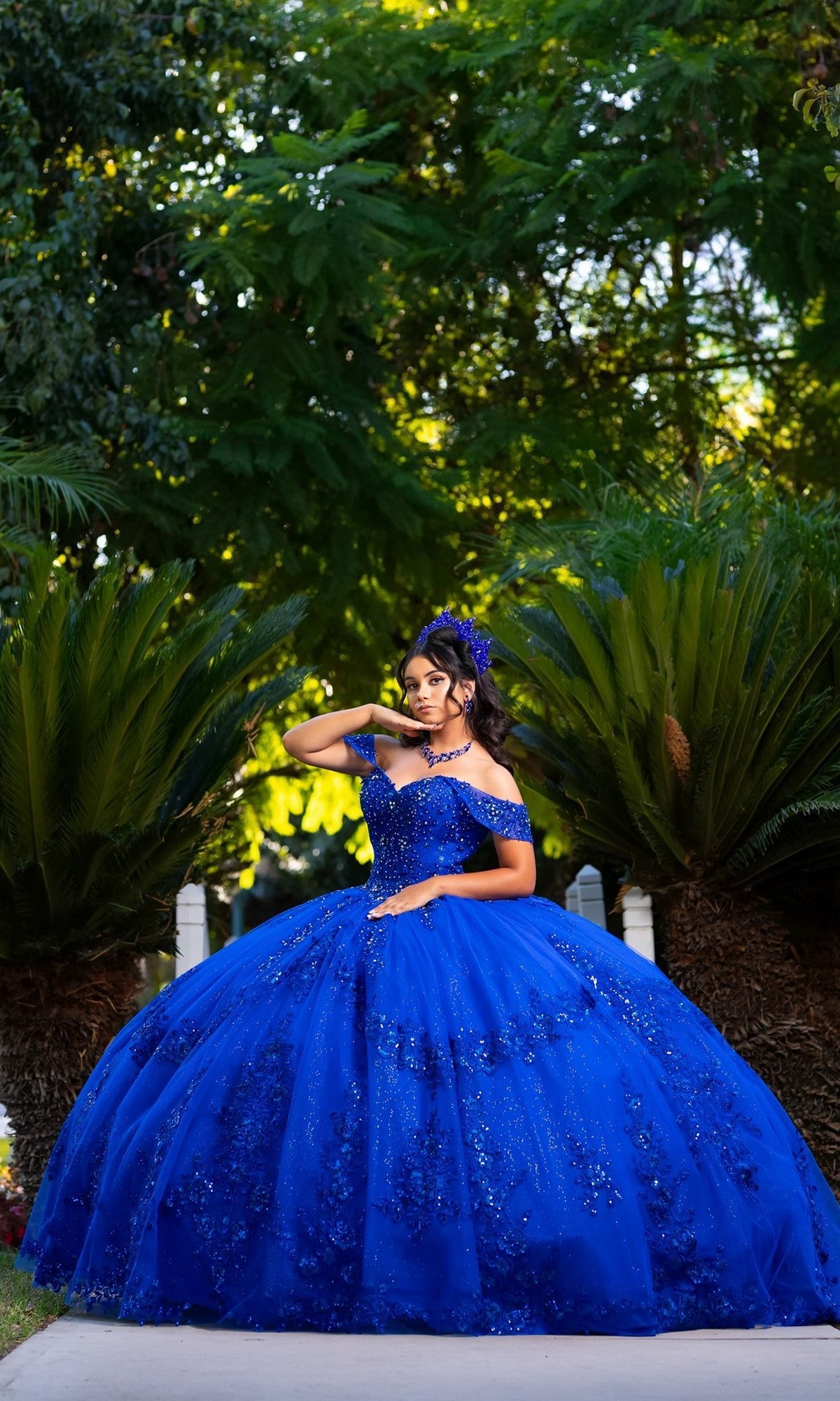 A young woman poses outdoors in the Quinceanera Dress CHF3122 by Chicas—a sparkling royal blue ballgown that beautifully complements the lush greenery around her. With one hand on her hip and the other touching her hair, she exudes grace in this elegant off-the-shoulder design, adding a sophisticated touch to the enchanting scene.